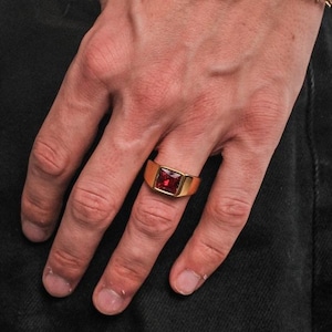 Puede incluir: Un anillo dorado con una gema roja cuadrada se lleva en un dedo. También se ve una pulsera dorada con un diseño de alambre de púas. La persona lleva vaqueros negros y una camiseta negra.