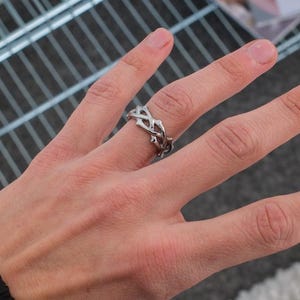 May include: A silver ring with a chain-like design. The ring is worn on the middle finger of a person's hand.