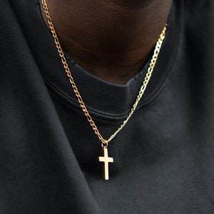 Puede incluir: Un collar de cadena de oro con un colgante de cruz de oro. El collar lo lleva una persona que lleva una camiseta negra.