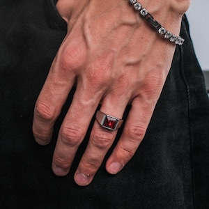 Könnte beinhalten: Ein silberner Ring mit einem rechteckigen, tiefroten Stein wird am Finger getragen. Ein Armband mit klaren Steinen und einem schwarzen Balken ist ebenfalls zu sehen. Die Person trägt eine schwarze Jeans und ein schwarzes Oberteil.