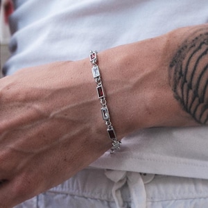 May include: A silver-toned chain bracelet featuring alternating rectangular baguette-cut stones in clear and deep red colors. The bracelet is constructed with small metal links connecting each stone setting, creating a flexible and delicate wrist accessory.