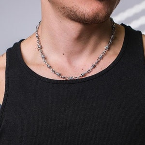 May include: A silver barbed wire chain necklace. The necklace is worn by a person with tattoos on their arms.