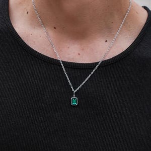 Puede incluir: Un collar de plata con un colgante rectangular verde esmeralda. El colgante está engastado en un marco de plata y cuelga de una delicada cadena de plata. La persona lleva una camiseta sin mangas negra.