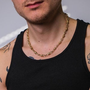 May include: A gold chain necklace with barbed wire links. The necklace is worn by a person with tattoos on their arms.