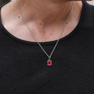 Könnte beinhalten: Eine silberne Halskette mit einem rechteckigen, roten Edelsteinanhänger. Der Anhänger ist in einen silbernen Rahmen gefasst und hängt an einer filigranen Silberkette. Die Person trägt ein schwarzes Tanktop.