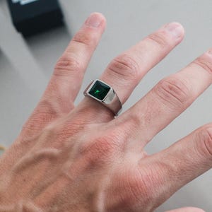 Könnte beinhalten: Ein silberner Ring mit einem quadratischen, smaragdgrünen Edelstein. Der Ring befindet sich an einem Finger, im Hintergrund sind weitere Ringe und Schachteln zu sehen. Der Ring hat ein modernes, minimalistisches Design.
