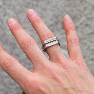 Könnte beinhalten: Ein silberner Ring mit zwei übereinanderliegenden rechteckigen Bändern. Der Ring wird am Zeigefinger einer Hand mit einer roten Tätowierung getragen, auf der "Immer" steht.