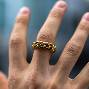 Può includere: Un anello a catena d'oro con un design spesso e massiccio. L'anello è indossato sul dito medio di una mano.