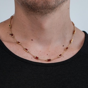 Könnte beinhalten: Eine goldfarbene Halskette mit rechteckigen, rotfarbenen Edelsteinen. Die Kette besteht aus kleinen goldenen Gliedern und wird um den Hals getragen. Der Hintergrund ist einfarbig hell.