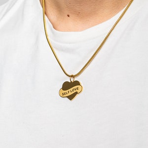 Könnte beinhalten: Eine goldene Kettenkette mit einem herzförmigen Anhänger, auf dem in schwarzen Buchstaben "SELF LOVE" steht.
