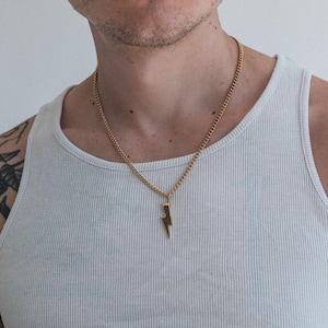 May include: Gold-colored necklace with a lightning bolt pendant. The chain is a curb style, and the pendant is a simple, stylized lightning bolt. The necklace is worn over a white ribbed tank top.