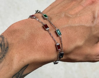 May include: Two silver chain bracelets with rectangular red and green gemstones. The bracelets are layered on a person's wrist.