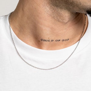 Pode incluir: Uma corrente de metal de tom prateado com um design de elos delicados. A corrente repousa sobre uma t-shirt branca lisa, destacando a textura metálica e a construção simples e clássica desta peça de joalharia à volta do pescoço.