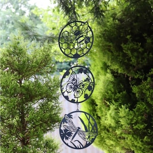 May include: A black metal garden ornament with three circular panels, each with a nature-themed cutout. The top panel features a bee and honeycomb, the middle a butterfly, and the bottom a dragonfly. The ornament is suspended by a chain.
