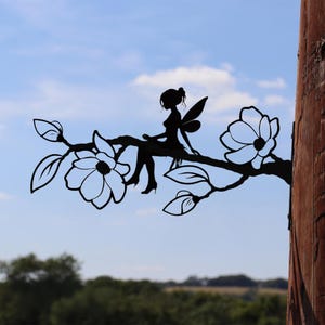 May include: Black metal silhouette of a fairy sitting on a branch with flowers and leaves. The fairy has wings and is wearing high heels. The branch is attached to a wooden post. The background is a blue sky with clouds.