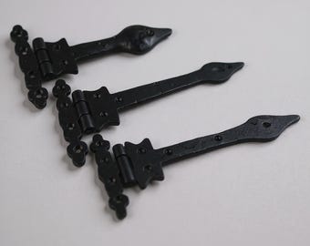 May include: Three black metal strap hinges with a decorative spear-shaped end and a textured finish. Each hinge features a rectangular mounting plate with multiple pre-drilled screw holes and a central barrel hinge mechanism for pivoting movement.