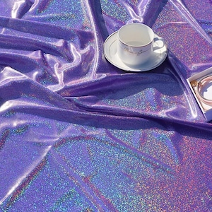 May include: A vibrant, iridescent purple fabric with a reflective surface. A white teacup and saucer with gold trim are on the fabric. A small white box with a pink bracelet is also present.
