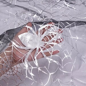 May include: Silver and black Halloween fabric with a spiderweb design. The fabric features a large silver spider and intricate web patterns, ideal for spooky decorations. The sheer material allows light to pass through, creating a ghostly effect.