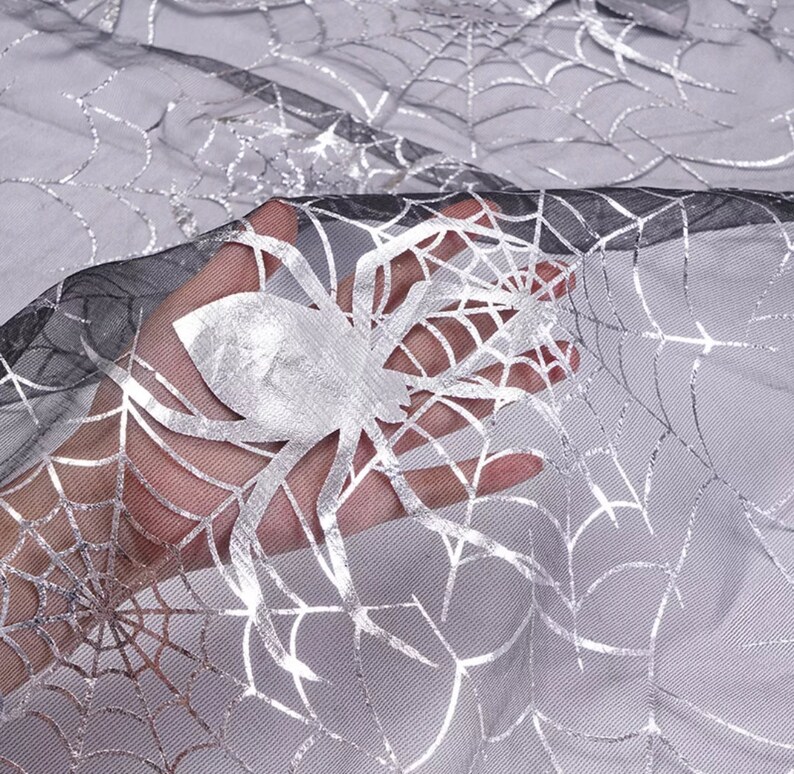 May include: Silver and black Halloween fabric with a spiderweb design. The fabric features a large silver spider and intricate web patterns, ideal for spooky decorations. The sheer material allows light to pass through, creating a ghostly effect.