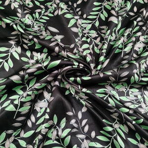 May include: Black satin fabric with a repeating pattern of green and white leaves and vines. The leaves have a detailed vein design. The fabric is swirled, creating shadows and highlights. The overall effect is a luxurious, botanical design.