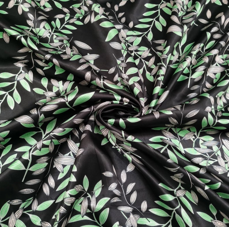 May include: Black satin fabric with a repeating pattern of green and white leaves and vines. The leaves have a detailed vein design. The fabric is swirled, creating shadows and highlights. The overall effect is a luxurious, botanical design.