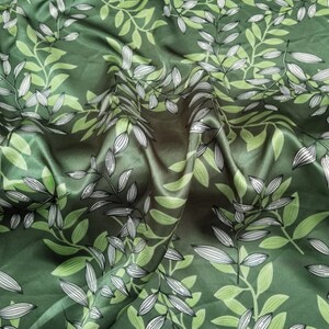 May include: Green fabric with a repeating pattern of stylized leaves and branches. The leaves are in shades of green and white, creating a natural, botanical design. The fabric has a slight sheen, suggesting a satin or silk material.