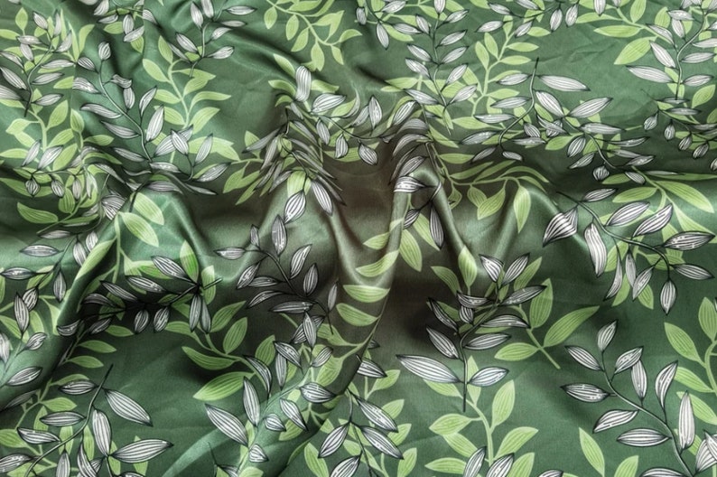 May include: Green fabric with a repeating pattern of stylized leaves and branches. The leaves are in shades of green and white, creating a natural, botanical design. The fabric has a slight sheen, suggesting a satin or silk material.