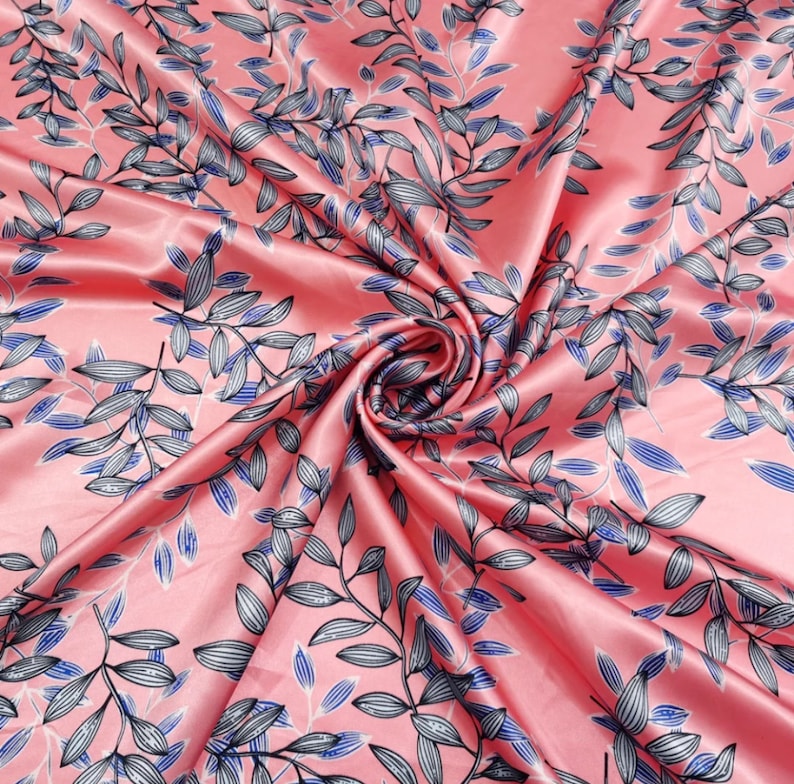 May include: A swirled, silky fabric with a pink background and a repeating pattern of stylized leaves. The leaves are primarily gray with blue accents and black outlines, creating a botanical design. The fabric has a glossy sheen.