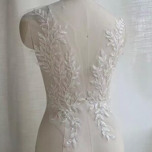 May include: White floral lace applique with a leaf design. The applique is made of delicate, shimmering fabric and is suitable for bridal or formal wear. The design features intricate detailing and a subtle, elegant aesthetic.