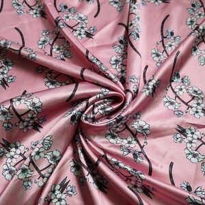 K&ouml;nnte beinhalten: Rosa Seidenstoff mit floralem Muster. Der Stoff zeigt wei&szlig;e Bl&uuml;ten mit schwarzen Stielen und &Auml;sten. Der Satin hat eine gl&auml;nzende Oberfl&auml;che und ist in weichen Wellen gefaltet, was einen luxuri&ouml;sen Look erzeugt.