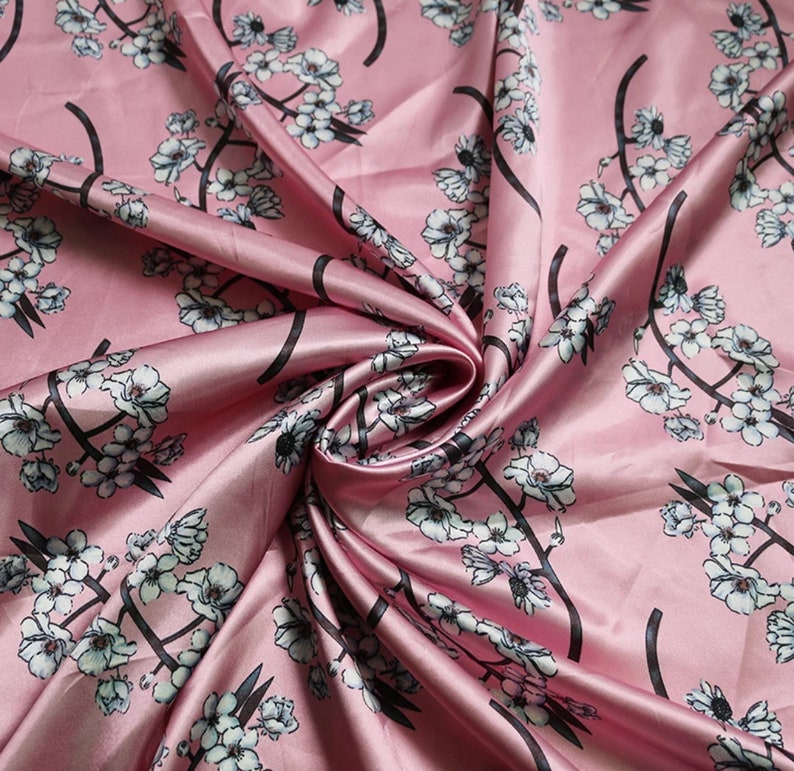 K&ouml;nnte beinhalten: Rosa Seidenstoff mit floralem Muster. Der Stoff zeigt wei&szlig;e Bl&uuml;ten mit schwarzen Stielen und &Auml;sten. Der Satin hat eine gl&auml;nzende Oberfl&auml;che und ist in weichen Wellen gefaltet, was einen luxuri&ouml;sen Look erzeugt.