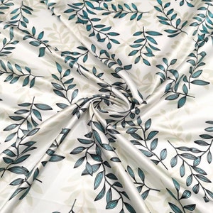 May include: White satin fabric with a repeating pattern of teal and black leaf branches. The fabric is folded and swirled, creating a dynamic display of the botanical design. The leaves have a detailed, hand-drawn appearance.