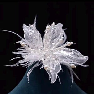 May include: A delicate, white floral hair accessory. The flower features layers of sheer fabric, embellished with sequins, pearls, and crystals. Feathery accents add texture. The design is intricate and elegant, suitable for weddings or special occasions.