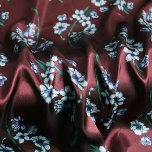 K&ouml;nnte beinhalten: Burgunderfarbener Satin-Stoff mit floralem Muster. Der Stoff zeigt wei&szlig;e und blaue Blumen mit gr&uuml;nen Stielen und Bl&auml;ttern. Das Material hat eine glatte, gl&auml;nzende Textur und ist in weichen Falten drapiert.