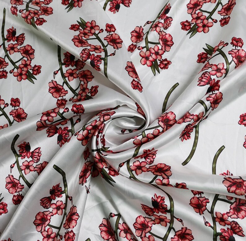 K&ouml;nnte beinhalten: Wei&szlig;er Satin-Stoff mit einem sich wiederholenden Muster aus roten und rosa Kirschbl&uuml;tenzweigen. Der Stoff ist drapiert und zeigt den Glanz und die Textur des Materials. Das florale Design ist detailliert und lebendig.