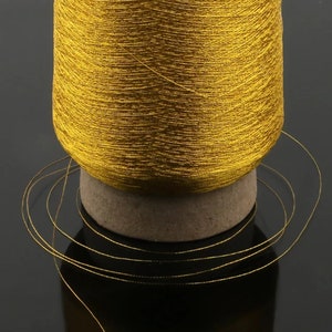 May include: A spool of golden-yellow thread, tightly wound, sits atop a small, cylindrical base. Several strands of the thread extend outwards, creating a delicate, looping pattern. The thread has a metallic sheen, reflecting light.