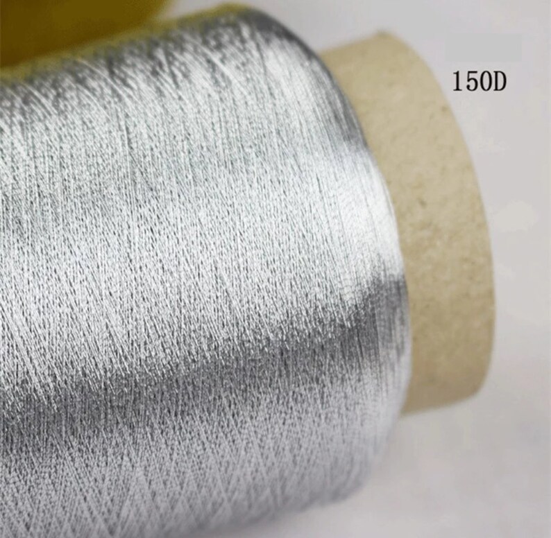 May include: A spool of silver-colored thread. The thread is tightly wound around a cardboard core. The text "150D" is printed on the side of the core. The thread has a metallic sheen.