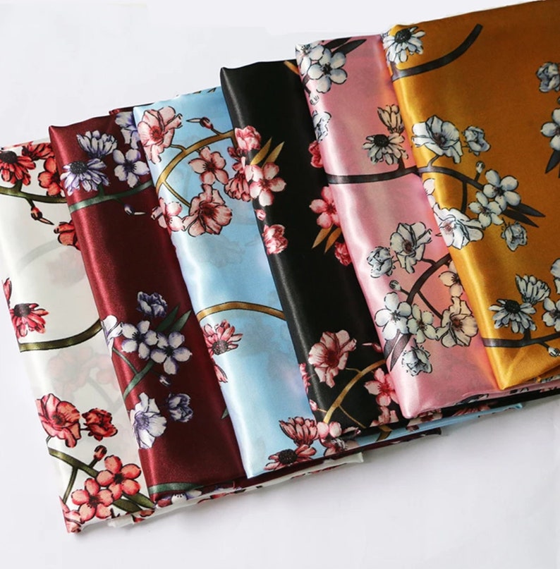 K&ouml;nnte beinhalten: Eine Sammlung von Seidenschals in verschiedenen Farben, jeweils mit einem floralen Muster verziert. Die Schals zeigen eine Reihe von Farbt&ouml;nen, darunter Wei&szlig;, Burgunderrot, Schwarz, Hellblau, Rosa und Gold. Das florale Design zeigt rosa und wei&szlig;e Bl&uuml;ten.