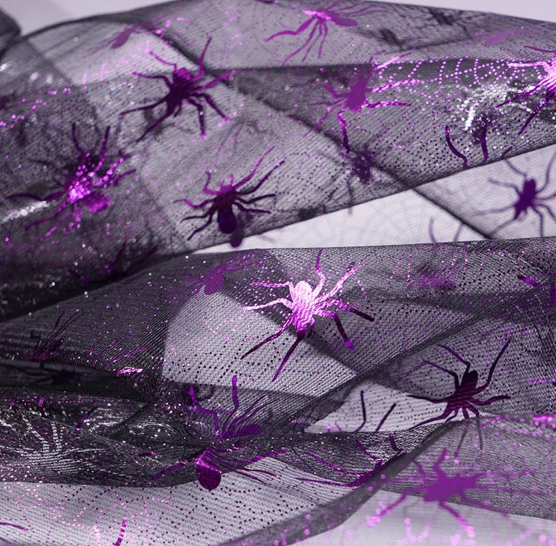May include: Black mesh fabric with glitter and purple spider accents. The spiders are in various sizes and poses, creating a spooky Halloween theme. The fabric is folded, showing the texture and design.