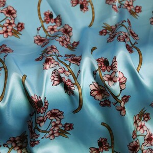 K&ouml;nnte beinhalten: Hellblauer Stoff mit einem sich wiederholenden Blumenmuster. Das Design zeigt rosa Bl&uuml;ten und braune Zweige vor einem weichen, flie&szlig;enden Hintergrund. Der Stoff hat einen seidigen Glanz.
