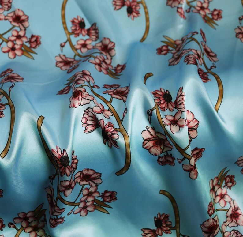 K&ouml;nnte beinhalten: Hellblauer Stoff mit einem sich wiederholenden Blumenmuster. Das Design zeigt rosa Bl&uuml;ten und braune Zweige vor einem weichen, flie&szlig;enden Hintergrund. Der Stoff hat einen seidigen Glanz.