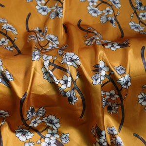 K&ouml;nnte beinhalten: Senffarbener Stoff mit floralem Muster. Das Design zeigt wei&szlig;e Bl&uuml;ten mit grauen Zentren und dunkelgrauen Zweigen. Der Stoff hat ein gl&auml;nzendes, satinartiges Aussehen mit sichtbaren Falten und Knitter.