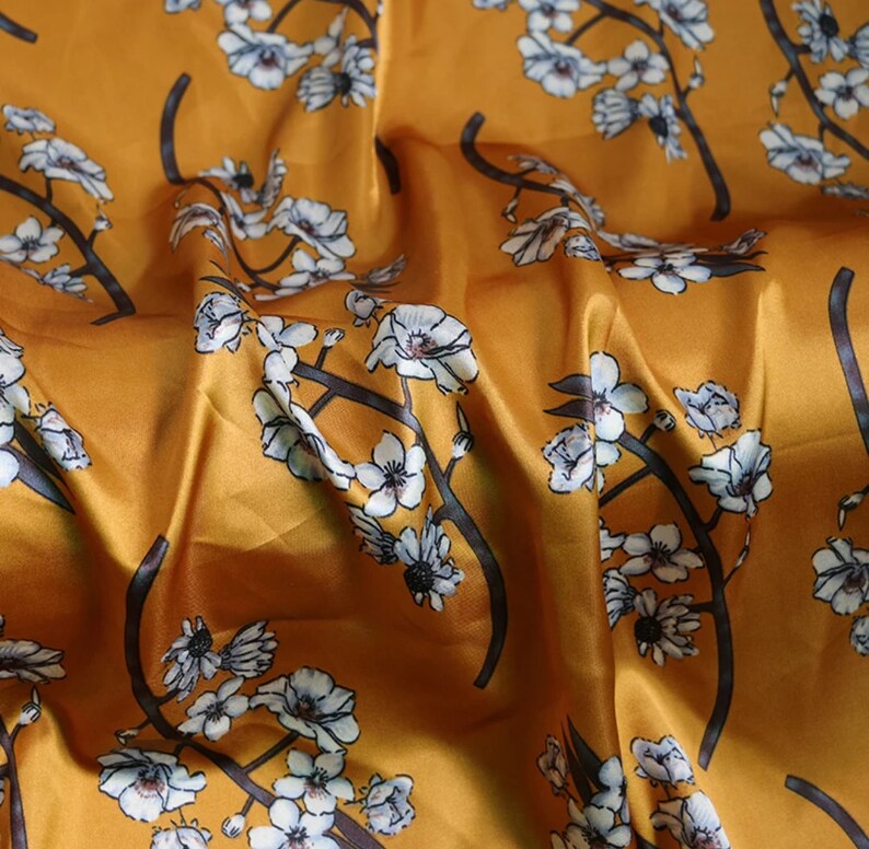 K&ouml;nnte beinhalten: Senffarbener Stoff mit floralem Muster. Das Design zeigt wei&szlig;e Bl&uuml;ten mit grauen Zentren und dunkelgrauen Zweigen. Der Stoff hat ein gl&auml;nzendes, satinartiges Aussehen mit sichtbaren Falten und Knitter.