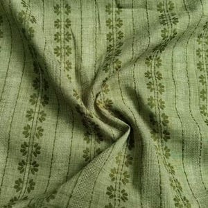 May include: Close-up of a green fabric with a floral pattern. The fabric has vertical stripes with a repeating floral design in a darker shade of green. The fabric is slightly wrinkled, showing its texture and drape.