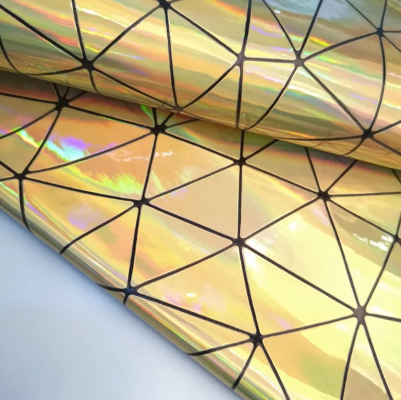May include: Close-up of a roll of iridescent fabric with a geometric pattern. The fabric has a gold and rainbow sheen, with black lines forming triangles and other shapes. The material appears shiny and reflective, suitable for crafts or fashion.