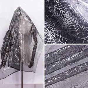 May include: Black, sheer fabric featuring a silver spiderweb design. The material is draped over a mannequin, highlighting the intricate pattern. Suitable for Halloween or themed occasions.