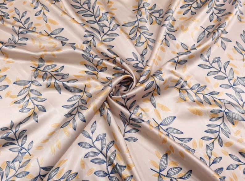 May include: Satin fabric with a repeating pattern of stylized leaves. The leaves are in shades of navy blue and gold against a pale beige background. The fabric is draped, showing folds and texture.
