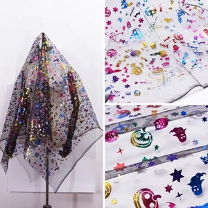May include: Black sheer fabric featuring a Halloween pattern. The fabric is decorated with colourful holographic images of stars, pumpkins, ghosts, and spiderwebs. The fabric is draped over a stand.