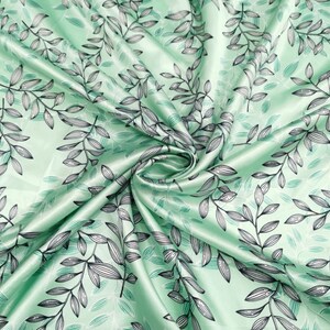 May include: A folded piece of fabric with a repeating pattern of black and gray leaves and stems on a light green background. The fabric has a satin sheen, and the leaves are detailed with fine lines.