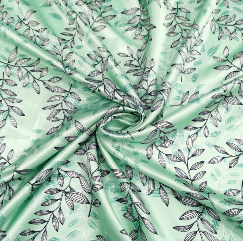 May include: A folded piece of fabric with a repeating pattern of black and gray leaves and stems on a light green background. The fabric has a satin sheen, and the leaves are detailed with fine lines.